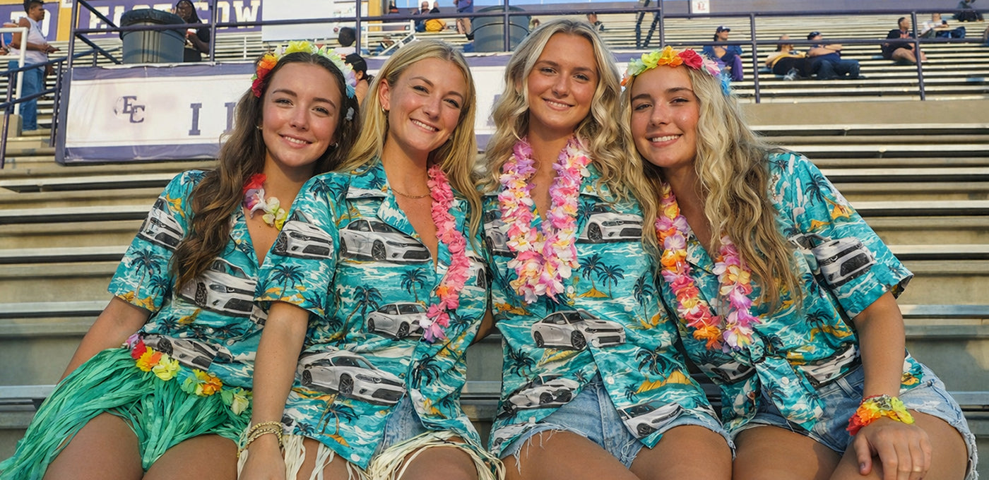 Four young women sitting on stadium bleachers, wearing matching car-print Hawaiian shirts and colorful leis, smiling together
