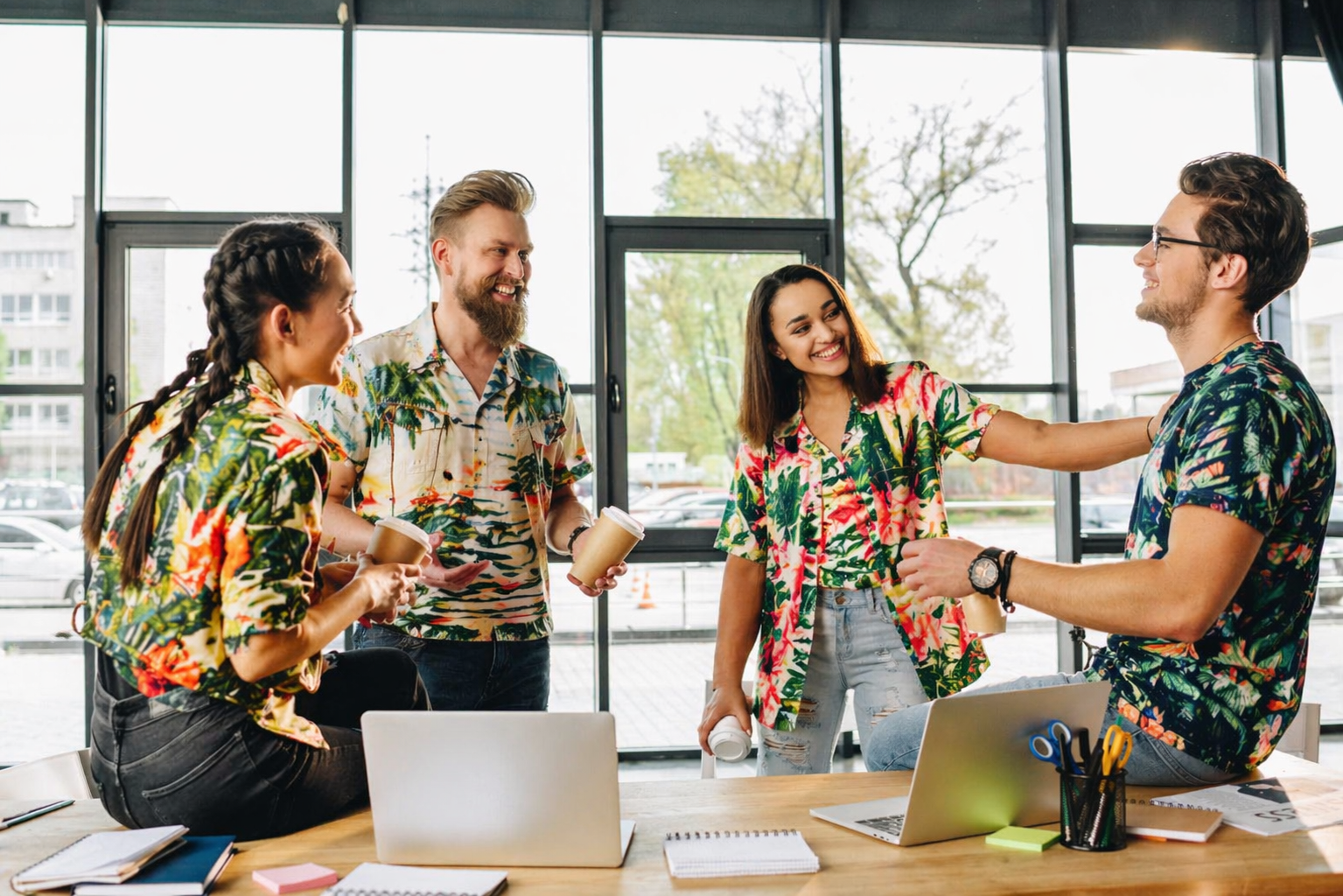 Team of coworkers in a modern office wearing tropical Hawaiian shirts, laughing and chatting during a casual coffee break