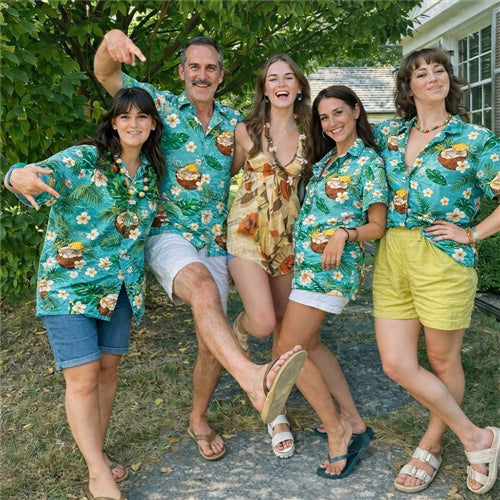 Five adults posing playfully outdoors, wearing matching green Hawaiian shirts with tropical coconut print